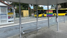 Eine totale Aufnahme von einer Bushaltestelle in Essen mit einem grauen Bushäuschen mit Glaspaneelen, im Hintergrund ist ein gelber Bus zu sehen.