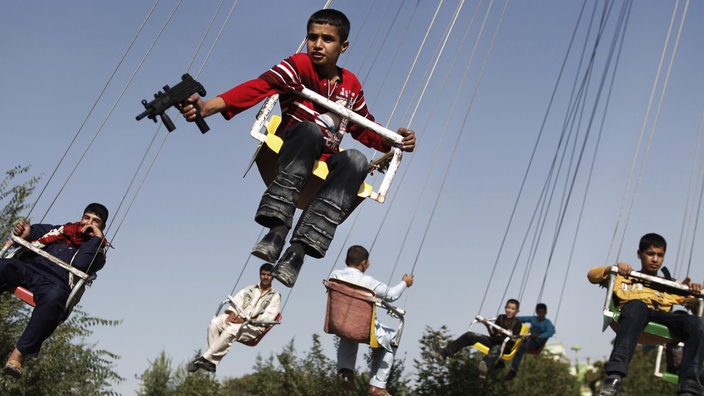 Mehrere afghanische Jungen sitzen auf einem Karussell, einer von ihnen hält eine Spielzeugwaffe