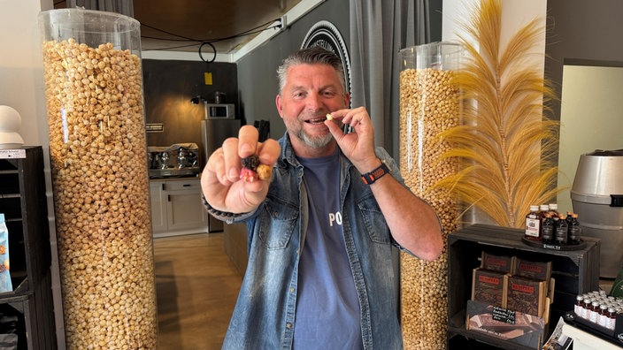 Michael Althoff hält mehrere Popcornstücke in die Kamera und isst eines, im Hintergrund große Behälter mit Popcorn.
