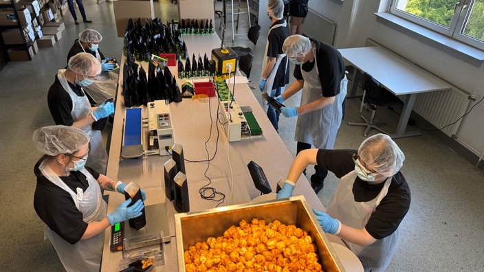 Mehrere Mitarbeitende mit Schutzkleidung füllen in einer Werkstatt große Mengen Popcorns ab.