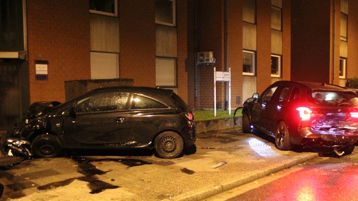 Zwei Autos mit Totalschaden stehen auf einem Bürgersteig vor einem Gebäude.