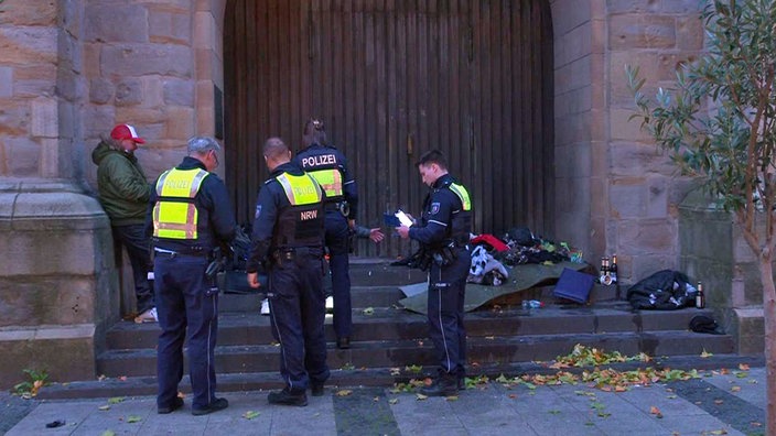 Polizisten vor  dem Portal einer Kirche mit Kleidung von Obdachlosen