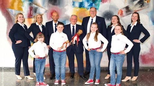 Mehrere Männer und Frauen in dunkelblauen Anzügen stehen lächelnd nebeneinander und schauen in die Kamera. Vor ihnen stehen vier Kinder in Jeans und weißen Hemden.