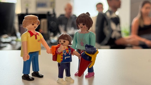 Auf einem Tisch stehen kleine Figuren einer Familie, im Hintergrund ist ein Büro mit Menschen.