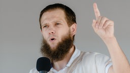 Junger Mann spricht mit erhobenem Zeigefinger in ein Mikrofon