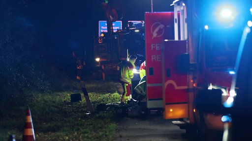 Rettungssanitäter knien an Unfallort. An der Seite stehen Rettungsfahrzeuge.Ein Baumfällfahrzeug ist im Hintergund zu sehen.