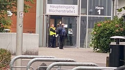 Vorm Haupteingang der Schule stehen zwei Personen in Warnweste und interagieren mit zwei weiteren Personen