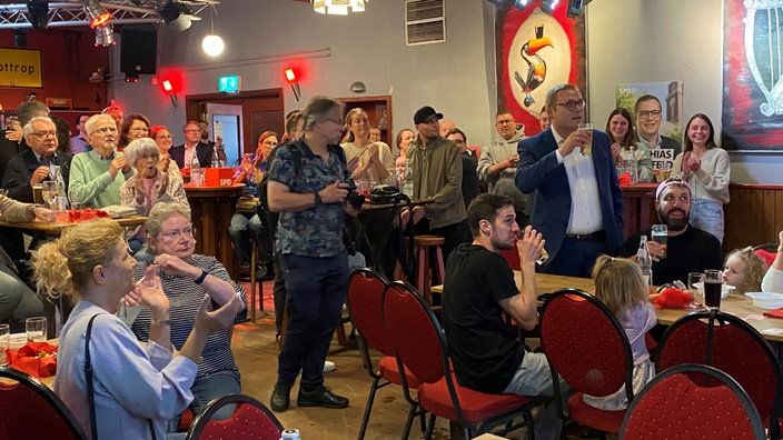 Meschen sitzen und stehen in einer Gaststätte, sie beobachten die Auszählung der Kommunalwahl