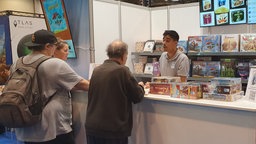 Robin Gupta steht hinter einem Spiele-Verkaufsstand und redet mit drei weiteren Personen