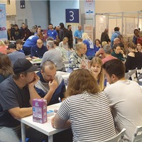 An mehreren Tischen in einer Halle sitzen Personen und spielen Spiele