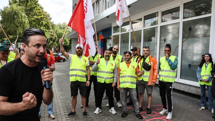Mehrere Menschen in gelben Warnwesten und mit Flaggen, im Vordergrund ein Mann mit Mikrofon
