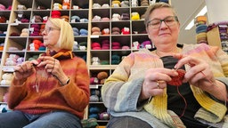 Zwei Frauen sitzen vor einem Regal voll Wolle und stricken