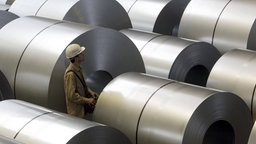 Auf dem Foto sind große Rollen feuerverzinkten Stahls. Zwischen den Rollen steht ein Arbeiter von Thyssenkrupp mit einem Helm.
