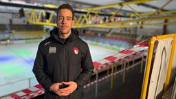 Ein Mann in schwarzer Jacke steht in der Duisburger Eishalle vor der Eisfläche und schaut in die Kamera.