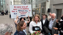 Das Bild zeigt eine Pressekonferenz. Die Anwältin des Kläger spricht, hinter ihr werden Protestschilder zur Klimagerechtigkeit hochgehalten.