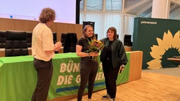 Katrin Lögering (mitte) wird ein Strauß von einer Person links von ihr überreicht. Eine weitere Person steht zu ihrer rechten Seite.