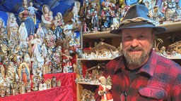 Ein Mann in Karohemd hält einen Weihnachtsmann aus Holz in der Hand, hinter ihm hunderte geschnitzte Figuren