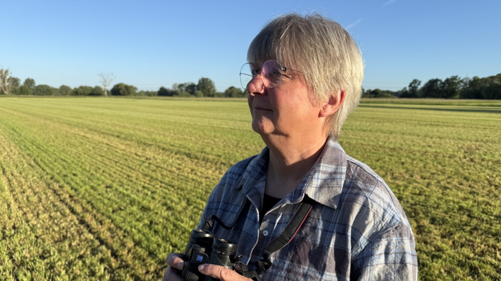 Eine Frau mit Brille steht auf einem Feld unf hält ein Fernglas in den Händen.