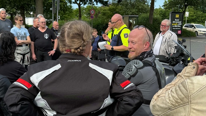 Menschen, einige in Motorradanzügen, umringen Polizisten, der etwas erklärt