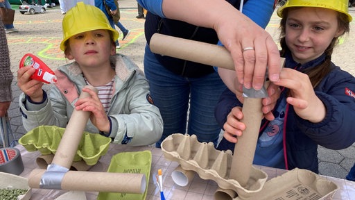 Zwei Kinder basteln mit Papprollen und tragen gelbe Bauhelme.
