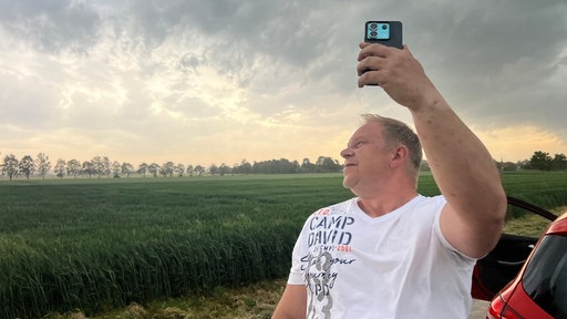 Sturmjäger André Ventrone hält sein Handy in die Luft und streamt für seine Follower. Er ist draußen, im Hintergrund sehen wir Gewitterwolken.