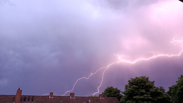Mehrere Blitze, die durch den lila, dunkel mit Wolken bedeckten Himmel fahren. Unten am Bild ist das Dach eines Hauses und ein Baum zu sehen.