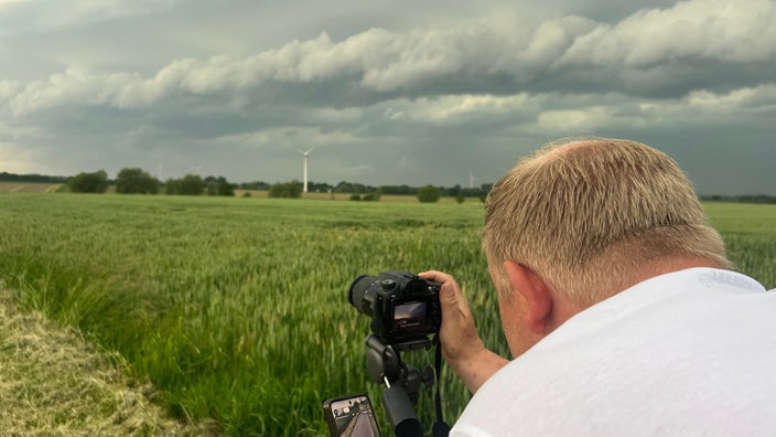 Sturmjäger André Ventrone steht hinter seiner Kamera und versucht das perfekte Bild zu machen vom Unwetter. Hinten am Horizont sind dunkle Wolen und viel Natur zu sehen.
