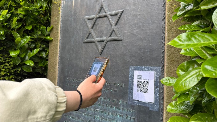 Eine Hand hält ein Smartphone auf einen QR-Code, der an der Gedenktafel zur abgebrannten Synagoge in Iserlohn klebt