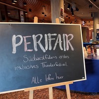 Im Eingangsbereich eines Gebäudes sind ein Tisch und mehrere Schilder mit der Aufschrift "Perifair" aufgestellt