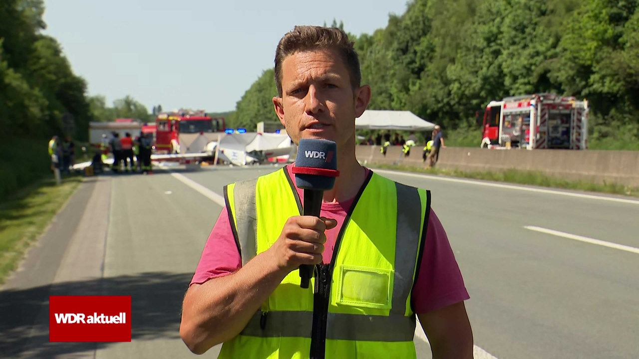 Ein Mann in Warnweste spricht in ein WDR-Mikrofon. Dahinter bergen Einsatzkräfte die Überreste eines Segelfliegers auf einer Autobahn.