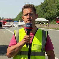 Ein Mann in Warnweste spricht in ein WDR-Mikrofon. Dahinter bergen Einsatzkräfte die Überreste eines Segelfliegers auf einer Autobahn.