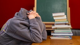 Eine Schülerin sitzt vor einem Stapel Büchern und hat trotzig die Kapuze ihres Pullovers über den Kopf gezogen
