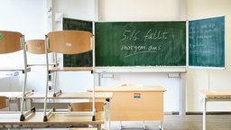 Ein leeres Klassenzimmer mit hochgestellten Stühlen und einer Tafel mit Schriftzug "5./6. fällt morgen aus!"