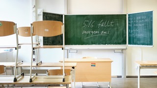 Ein leeres Klassenzimmer mit hochgestellten Stühlen und einer Tafel mit Schriftzug "5./6. fällt morgen aus!"