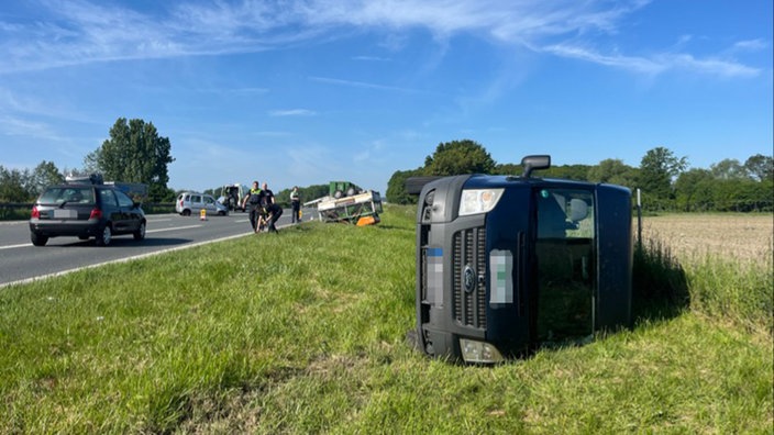 Ein schwarzer Transporter liegt seitlich auf der Wiese neben der Fahrbahn. Im Hintergrund sind ein Anhänger und zwei weitere Fahrzeuge.