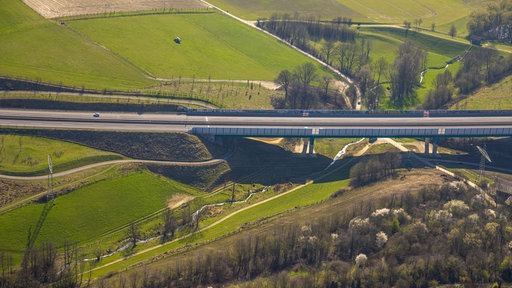 Eine Autobahnbrücke führt über eine grüne Landschaft mit Feldern, Bäumen und einem kleinen Tal darunter.