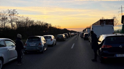 Autos stehen bei Sonnenuntergang dicht hintereinander auf einer Autobahn, Menschen stehen neben den Fahrzeugen.
