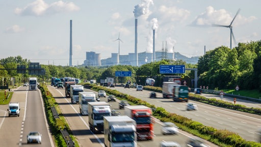 Viele LKW fahren auf der Autobahn A2, hinten das Uniper Steinkohlekraftwerk Gelsenkirchen Scholven, rechts ein Windrad.
