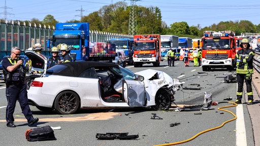 Ein stark beschdigtes, weißes Auto auf der autobahn, drumherum Rettungwagen und Polizei