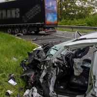 Ein stark beschädigtes weißes Fahrzeug steht an einem Straßenrand. Auf der Straße steht ein blauer LKW sowie herumliegende Fahrzeugteile.