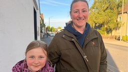 Eine Frau und ein junges Mädchen stehen auf den Gehweg einer Straße und lächeln in die Kamera