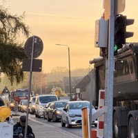 Eine dicht befahrende Straße, am Straßenrand stehen Baustellenschilder. Im Hintergrund orangefarbener Himmel. 