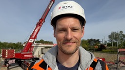 Ein Mann mit weißem Sicherheitshelm lächelt in die Kamera. Im Hintergrund ein roter Baukram. 