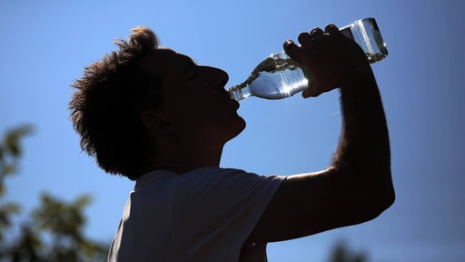 Silhouette eines Mannes, der aus einer Wasserflasche trinkt, im Hintergrund blauer Himmel