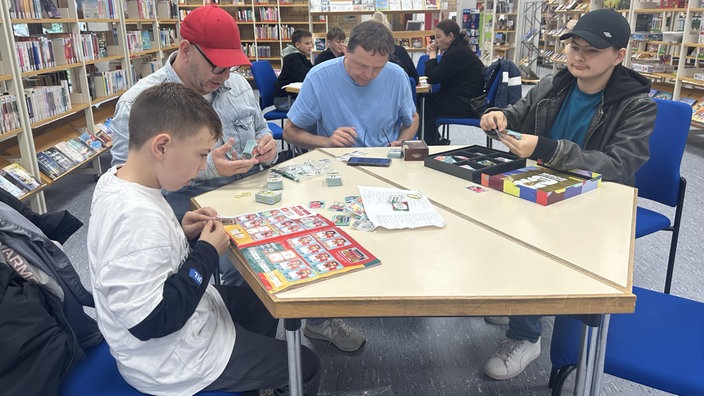 Vier Menschen sitzen an einem Tisch in der Stadtbibliothek Duisburg. Links ein Junge, daneben ein Mann mit roter Kappe, daneben ein Mann mit blauen T-Shirt und ganz rechts noch ein Junge. Auf dem Tisch liegen Sticker.