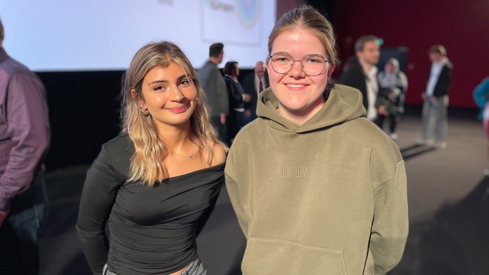 Melek Günes und Jule Stenzel stehen vor der Kinoleinwand und lächeln in die Kamera.