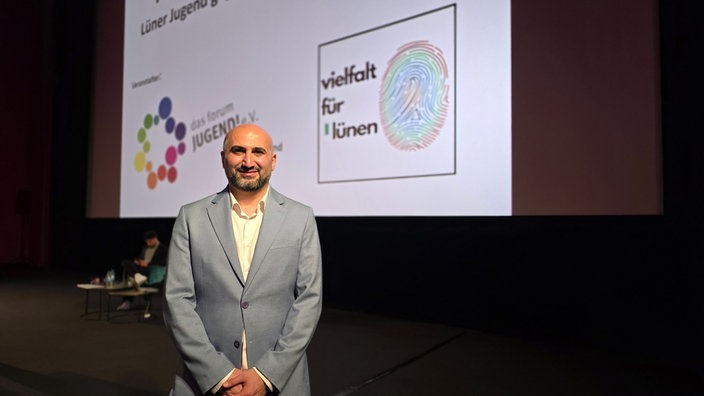 Ümithan Yağmur steht vor der Kinoleinwand mit den Logos vom "Forum Jugend" und der Wahlliste "Vielfalt für Lünen".