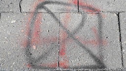 Auf einem Gehweg wurde mit roter Farbe ein Hakenkreuz gemalt, dieses wird von einem Kreuz in schwarzer Farbe durchgestrichen