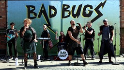 Die Inszenierung Bad Bugs des Theater Marabu in Bonn. Auf der Bühne stehen Menschen in kostümen, Sie singen in Mikrofone und spielen Instrumente. Im Hintergrund steht Bad Bugs auf einer Wand.