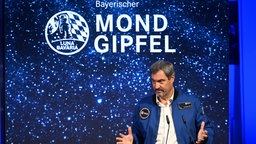 Der Bayerische Ministerpräsident Markus Söder (CSU) trägt eine Astronautenjacke, im Hintergrund ist das bayerische Landeswappen und der Schriftzug "Mondgipfel"